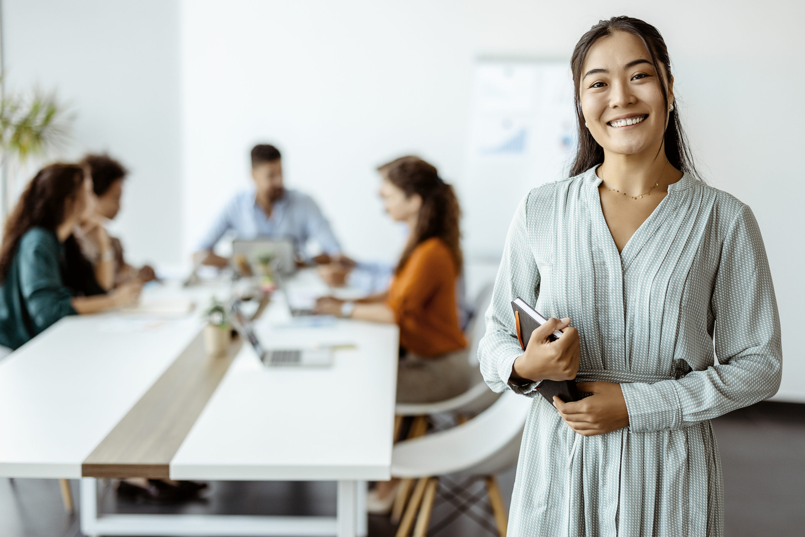 Confident young business woman standing smiling at the camera in a boardroom with colleagues in the background