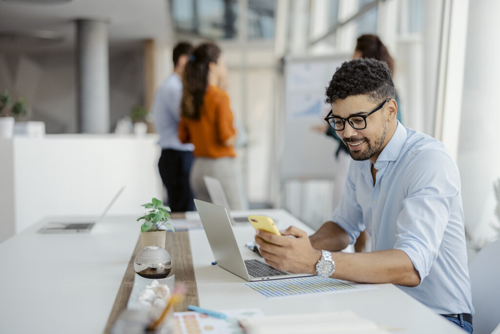 Happy african american businessman using phone mobile corporate apps at workplace texting sms, smiling black man looking at smartphone browsing internet, office technology and digital communication with colleagues in the background.